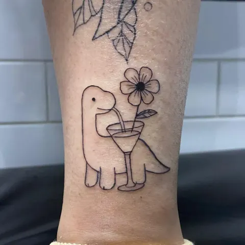Dinosaur with a cocktail and flower tattoo on the lower leg, by Kayla Grimwood