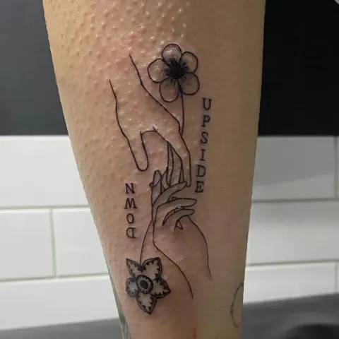 Reaching hands with a flower and Upside Down lettering tattoo on the forearm, by Kayla Grimwood