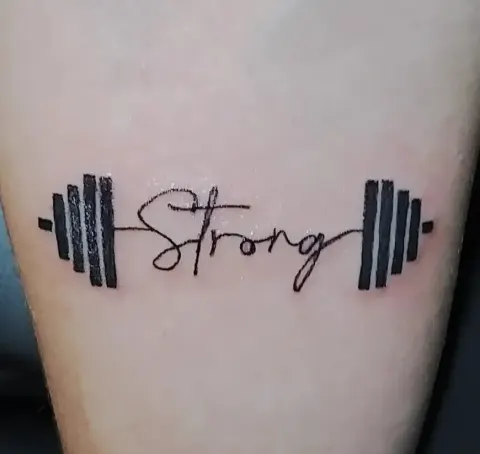 Strong lettering between dumbbell plates tattoo, by Kayla Grimwood