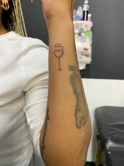 Wine glass with a heart and halo tattoo on the forearm, by Kayla Grimwood