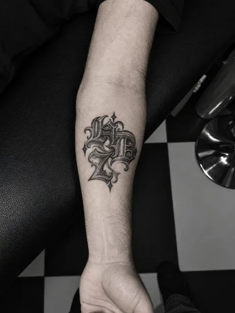 Black and grey gothic lettering monogram with sparkles on the forearm, by Rob Turner