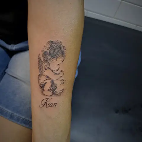 Cherub with a name tattoo on the forearm, by Tayla Symons