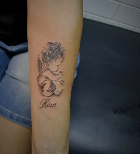 Cherub with a name tattoo on the forearm, by Tayla Symons