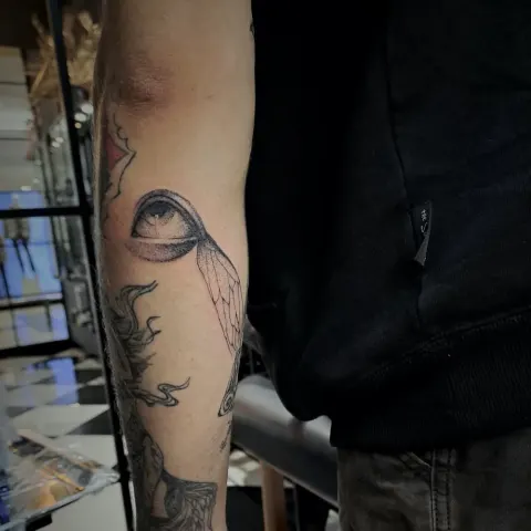 Upward-looking eye with a single wing, dotwork forearm tattoo, by Tayla Symons